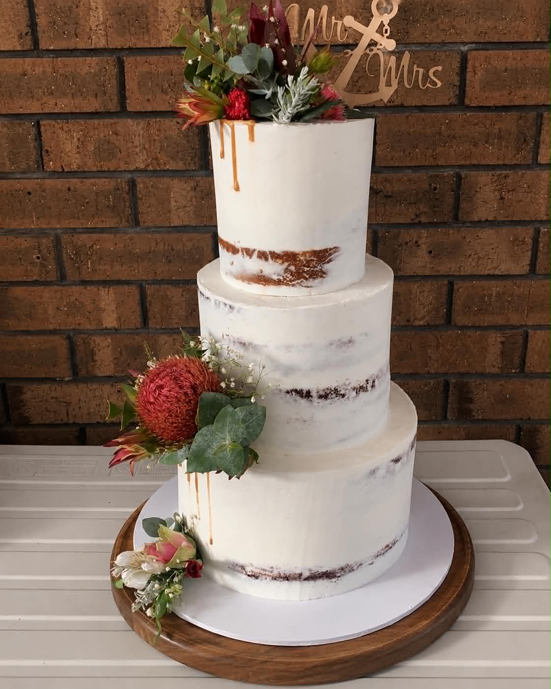 Three tier semi-naked wedding cake with caramel drip, red pincushion protea, pink and white flowers, eucalyptus and a gold Mr & Mrs acrylic topper on a wooden cake board